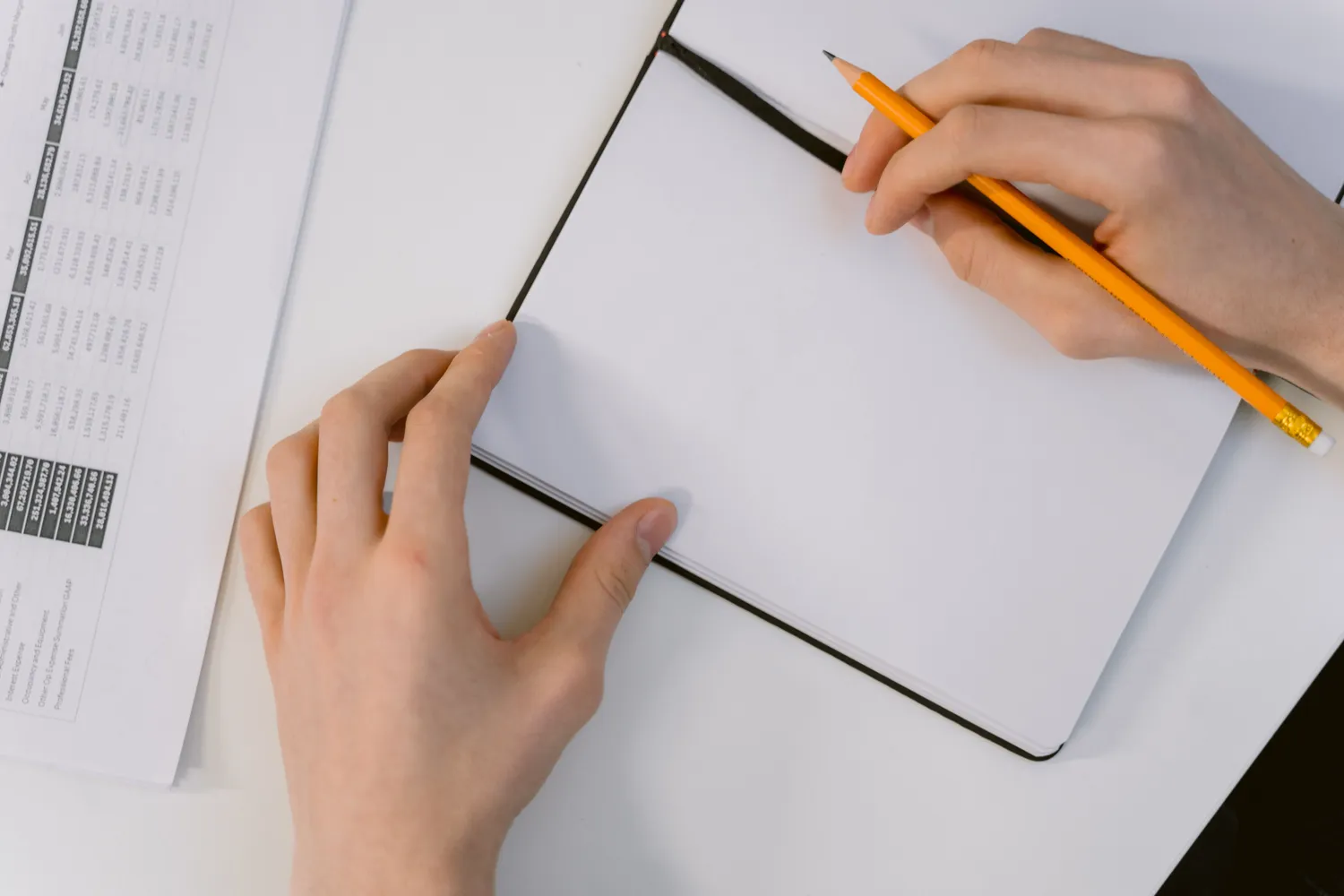 Hands holding a pencil over an open blank notebook on a light desk.